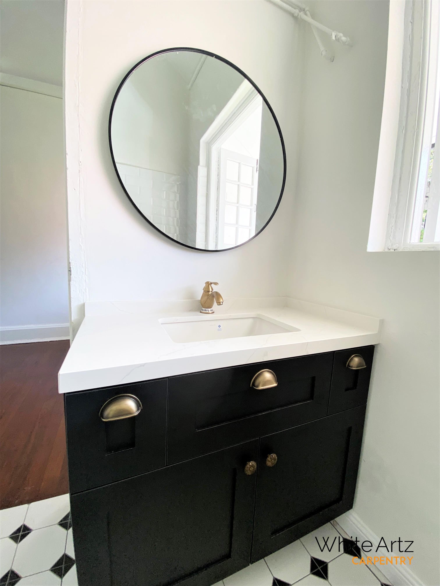 Custom black Shaker bathroom vanity cabinet by White Artz Carpentry, featuring a white quartz top, antique brass cup pulls, and a circular mirror.